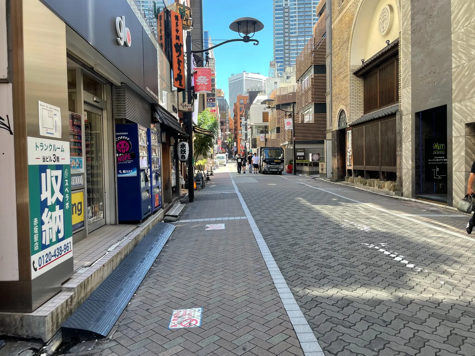 車道と歩道が同じ石張りで舗装された通り。左に自動販売機と「トランクルーム」の看板、右に木の格子を用いた建物