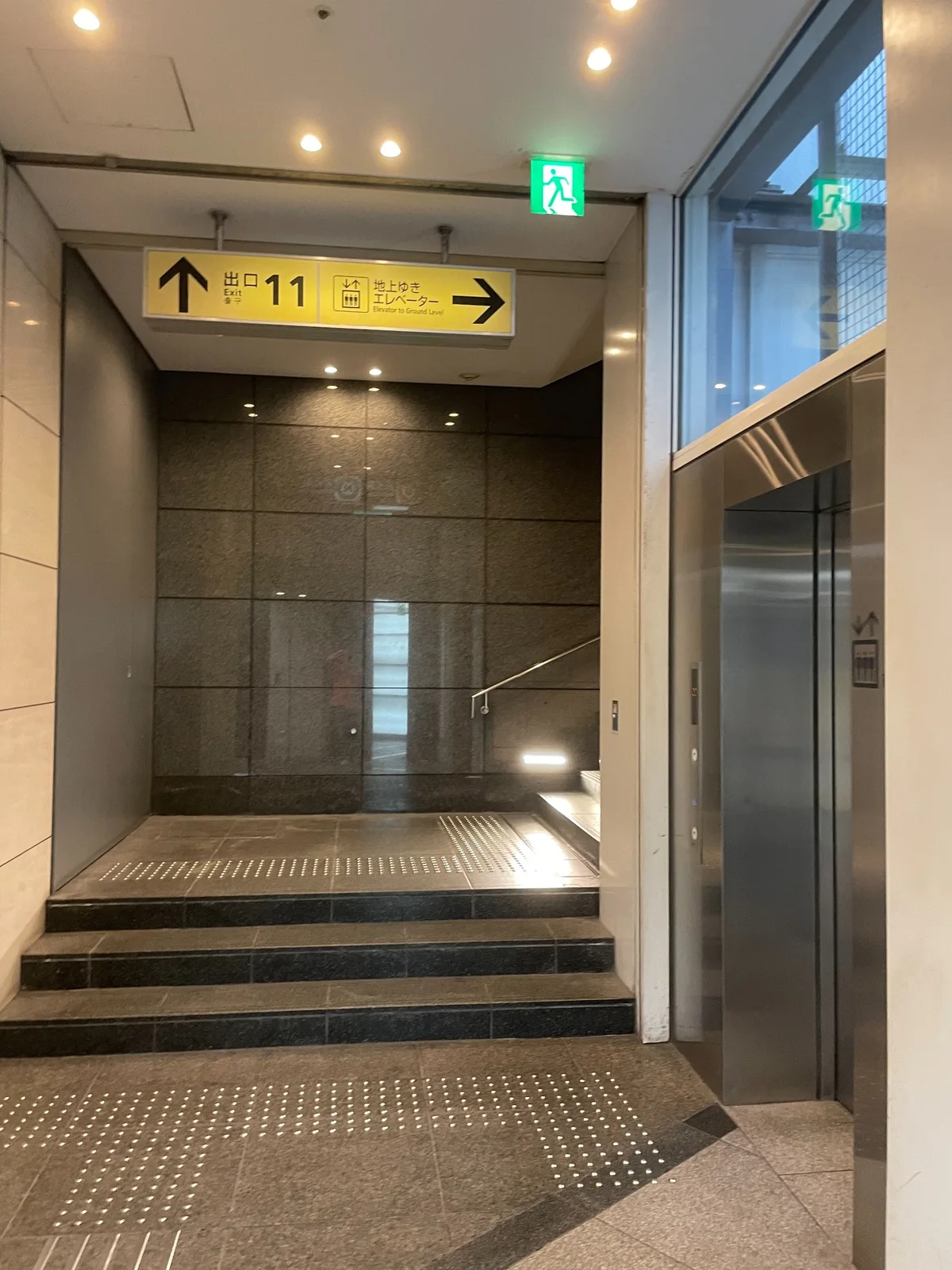 駅構内の黄色い案内看板。左に上向き矢印と「出口 11」、右に「地上ゆき エレベーター」